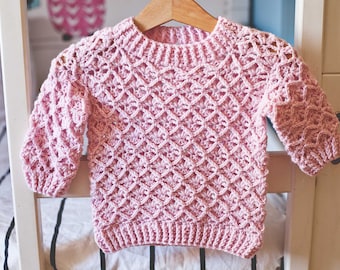 May include: A pink crocheted sweater with a diamond pattern. The sweater has a round neckline and long sleeves.