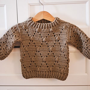 May include: A brown crocheted sweater with a round neckline and long sleeves. The sweater has a textured pattern with small holes throughout.