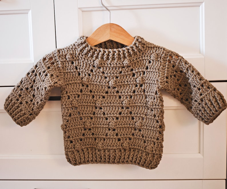 May include: A brown crocheted sweater with a round neckline and long sleeves. The sweater has a textured pattern with small holes throughout.