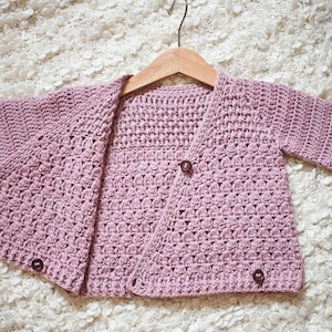 May include: A light purple crocheted cardigan with a button closure. The cardigan is hanging on a wooden hanger.