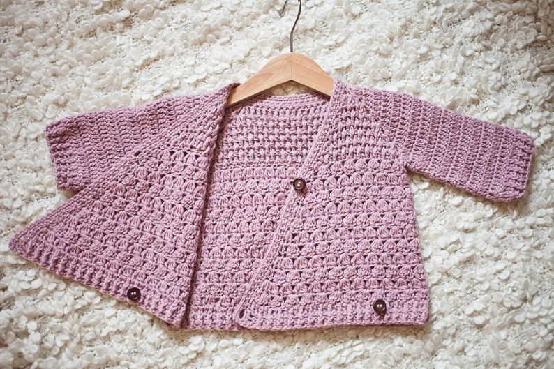 May include: A light purple crocheted cardigan with a button closure. The cardigan is hanging on a wooden hanger.