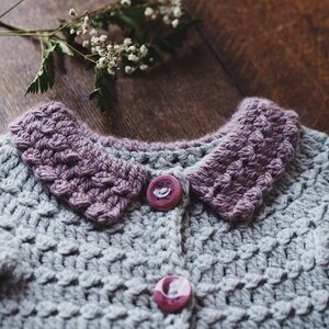 May include: A handmade gray crochet cardigan with a textured stitch pattern. The cardigan features a lavender collar and two matching buttons. A sprig of white flowers rests on a wooden surface near the garment.