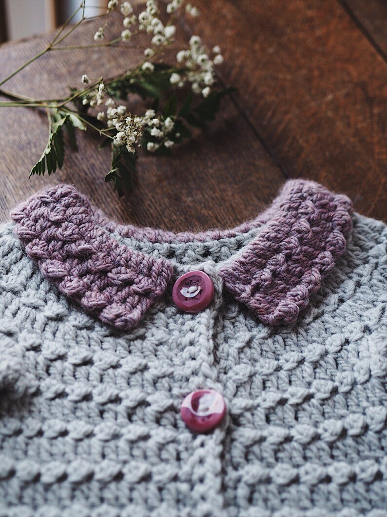 May include: A handmade gray crochet cardigan with a textured stitch pattern. The cardigan features a lavender collar and two matching buttons. A sprig of white flowers rests on a wooden surface near the garment.
