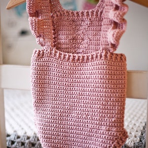 May include: A pink crocheted baby onesie with ruffled straps hanging on a wooden hanger.