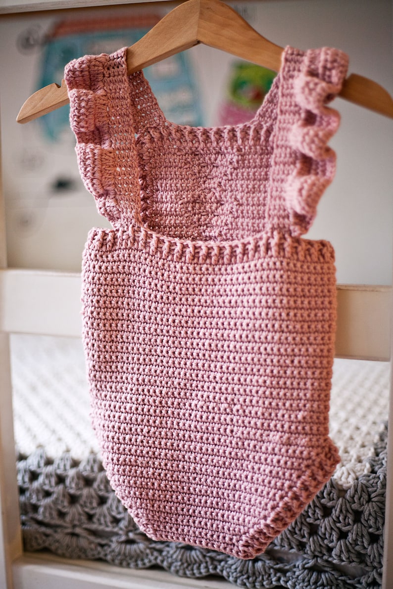 May include: A pink crocheted baby onesie with ruffled straps hanging on a wooden hanger.