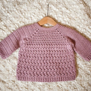 May include: A soft, pink crocheted sweater with a round neckline and long sleeves. The sweater is hanging on a wooden hanger.