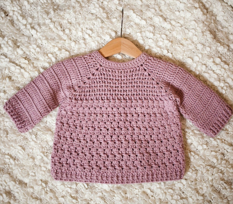 May include: A soft, pink crocheted sweater with a round neckline and long sleeves. The sweater is hanging on a wooden hanger.