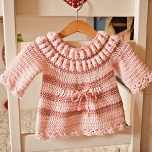 May include: A pink crocheted baby sweater with a ruffled neckline and a tie at the waist.