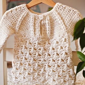 May include: A white crocheted baby sweater with a delicate openwork pattern. The sweater has a round neckline and long sleeves.