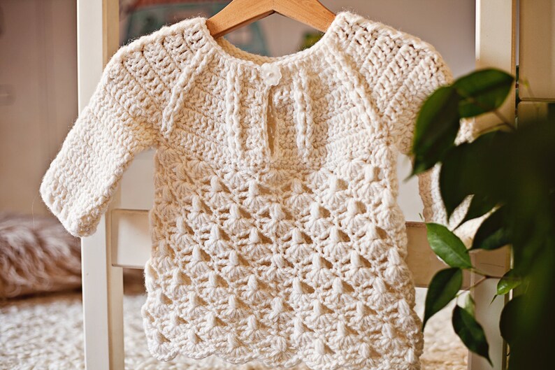 May include: A white crocheted baby sweater with a delicate openwork pattern. The sweater has a round neckline and long sleeves.