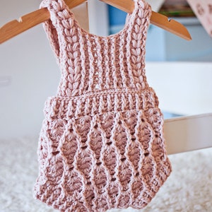 Op de afbeelding: Een roze gebreide baby romper met een delicaat, kantpatroon. De romper is mouwloos en heeft een gegolfde zoom.