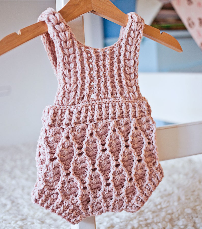 Op de afbeelding: Een roze gebreide baby romper met een delicaat, kantpatroon. De romper is mouwloos en heeft een gegolfde zoom.
