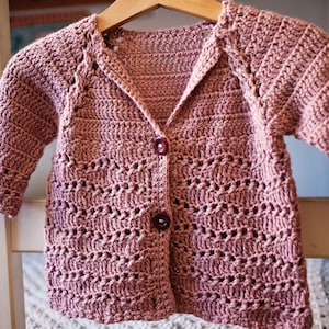 May include: A crocheted cardigan in a dusty rose color with a button closure. The cardigan has a delicate open stitch pattern and a slightly oversized fit.