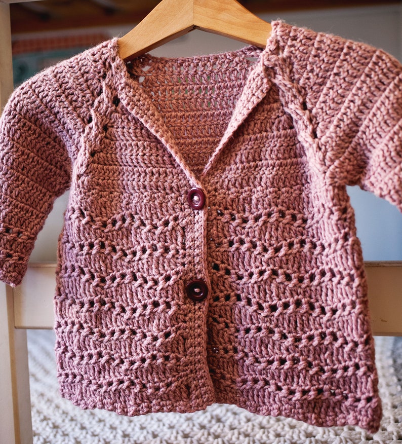 May include: A crocheted cardigan in a dusty rose color with a button closure. The cardigan has a delicate open stitch pattern and a slightly oversized fit.