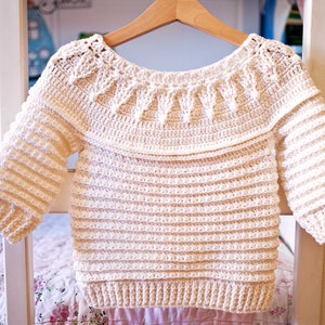 May include: Cream-colored, handmade crochet sweater with long sleeves. The sweater features a decorative neckline and is displayed on a wooden hanger. The crochet pattern is visible throughout the garment, showcasing the texture and craftsmanship.