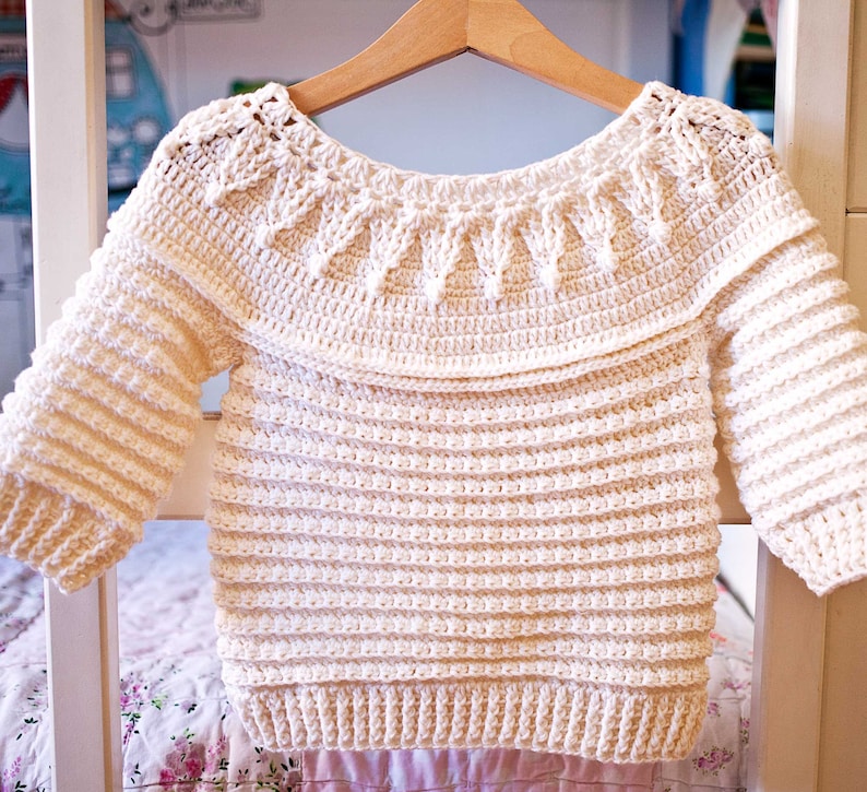 May include: Cream-colored, handmade crochet sweater with long sleeves. The sweater features a decorative neckline and is displayed on a wooden hanger. The crochet pattern is visible throughout the garment, showcasing the texture and craftsmanship.