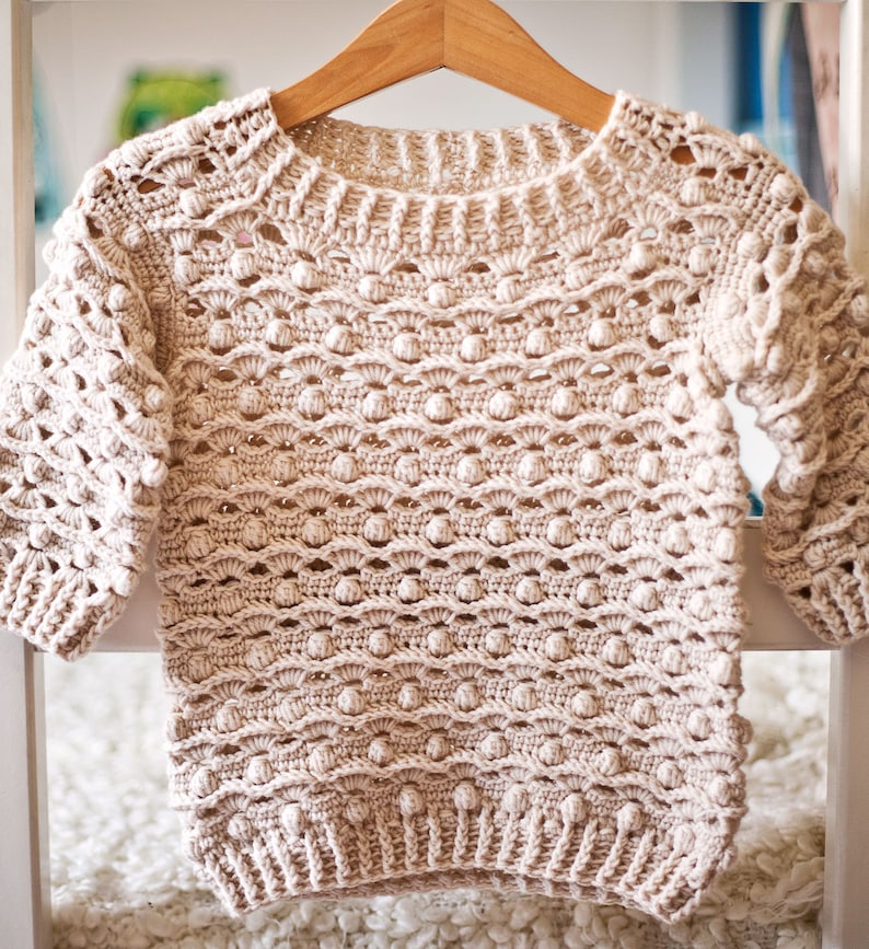May include: A beige crocheted sweater with a textured, bobble stitch pattern. The sweater has a round neckline and three-quarter sleeves.