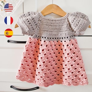 May include: A handmade crochet dress in gray and pink, displayed on a wooden hanger. The top is gray with short sleeves, and the skirt is a scalloped pink. The dress is likely for a baby or toddler.