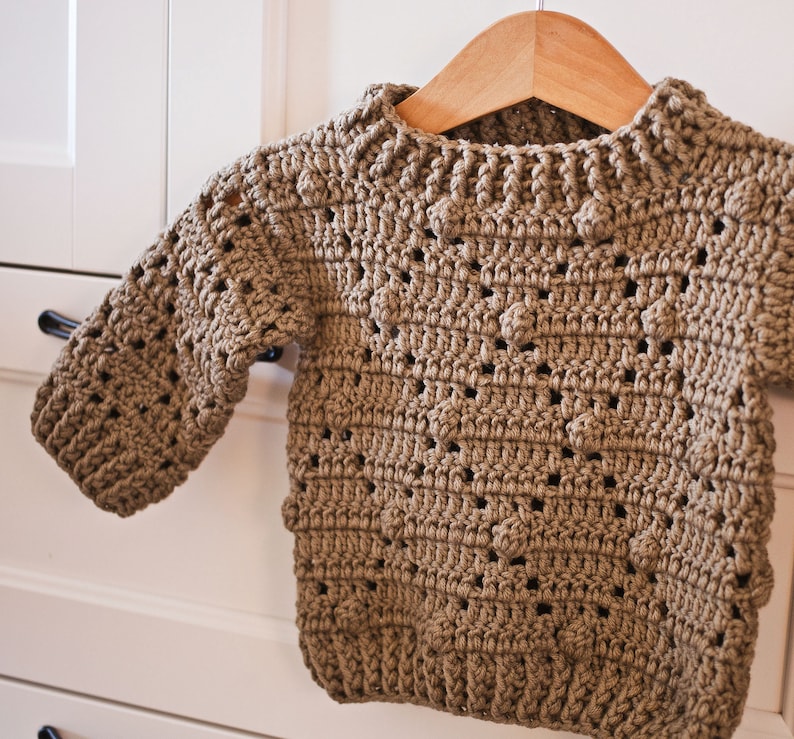 May include: A brown crocheted sweater with a textured pattern. The sweater has a round neckline and long sleeves.