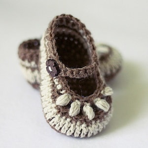 May include: A pair of crocheted baby shoes in brown and white with a brown button. The shoes have a scalloped edge and a small ruffle around the ankle.