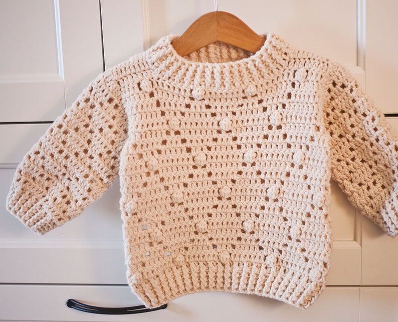 May include: A crocheted cream-colored sweater with a textured pattern. The sweater has long sleeves and a round neckline.
