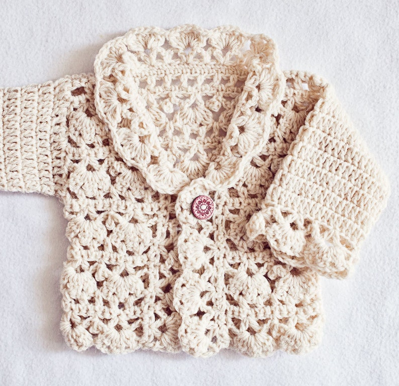 May include: Cream-colored, crocheted baby cardigan with a scalloped collar and cuffs. The button is a round, reddish-brown color. The crochet pattern is intricate, with a floral design. The cardigan is made of soft yarn.