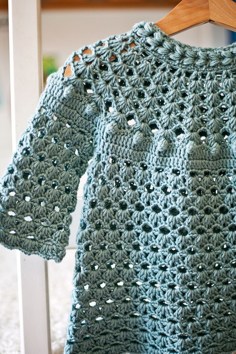 Puede incluir: Un vestido de beb&eacute; de ganchillo azul claro con un delicado patr&oacute;n de calado y peque&ntilde;as bolitas de ganchillo. El vestido tiene un escote redondo y mangas largas.