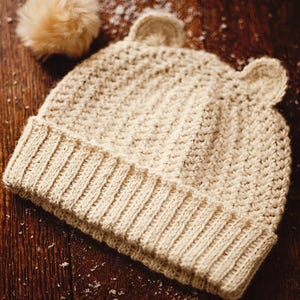 May include: A cream knitted beanie with ear flaps. The beanie has a textured stitch pattern and a fold-over brim.