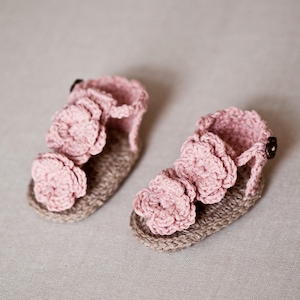 May include: A pair of pink crocheted baby sandals with flower embellishments. The sandals are made with a brown jute sole.