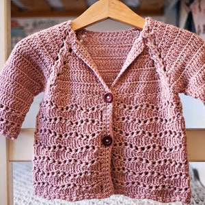 May include: A pink crocheted cardigan with a button fastening and a V-neck. The cardigan features a delicate openwork pattern.