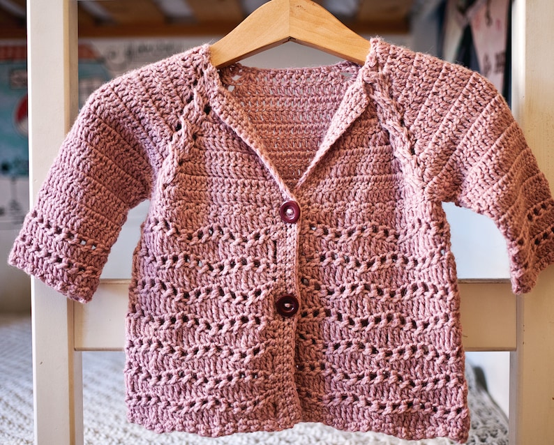 May include: A pink crocheted cardigan with a button closure and a V-neck. The cardigan has a delicate openwork pattern.
