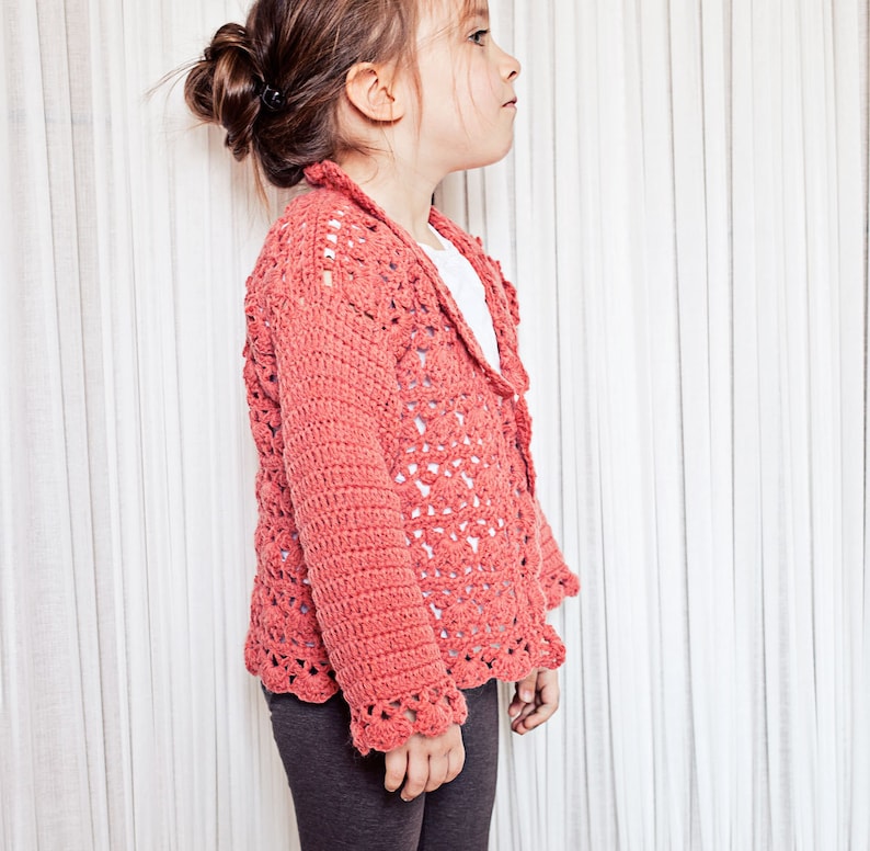 May include: Coral-colored crochet cardigan with a scalloped hem and openwork design. The long-sleeved sweater has a collar and is worn over a white top. The cardigan is made of a soft yarn and is suitable for children.
