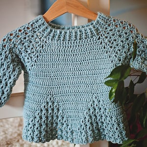 K&ouml;nnte beinhalten: Hellblauer, handgeh&auml;kelter Pullover f&uuml;r ein Baby oder Kleinkind. Der Pullover hat lange &Auml;rmel und einen runden Halsausschnitt. Das H&auml;kelmuster umfasst Lochmusterdetails an den &Auml;rmeln und am Passe. Das Kleidungsst&uuml;ck wird auf einem Holzb&uuml;gel pr&auml;sentiert.