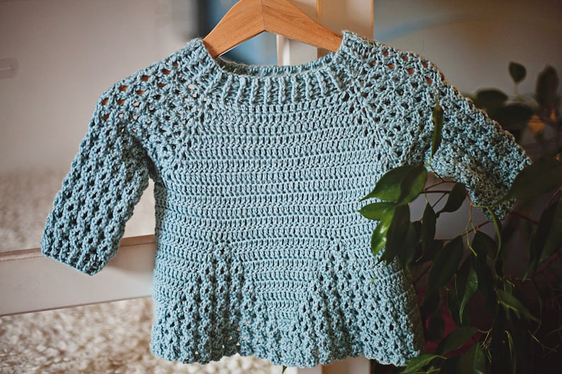 K&ouml;nnte beinhalten: Hellblauer, handgeh&auml;kelter Pullover f&uuml;r ein Baby oder Kleinkind. Der Pullover hat lange &Auml;rmel und einen runden Halsausschnitt. Das H&auml;kelmuster umfasst Lochmusterdetails an den &Auml;rmeln und am Passe. Das Kleidungsst&uuml;ck wird auf einem Holzb&uuml;gel pr&auml;sentiert.