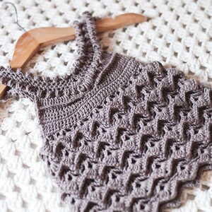May include: A crocheted sleeveless top in a light brown colour with a delicate stitch pattern. The top is hanging on a wooden hanger.