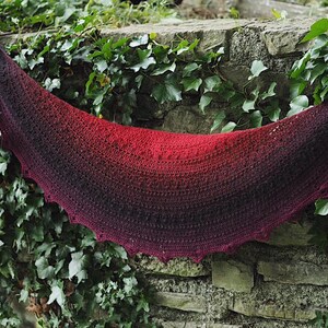May include: A crocheted shawl with a gradient of red to black. The shawl is draped over a stone wall covered in ivy.
