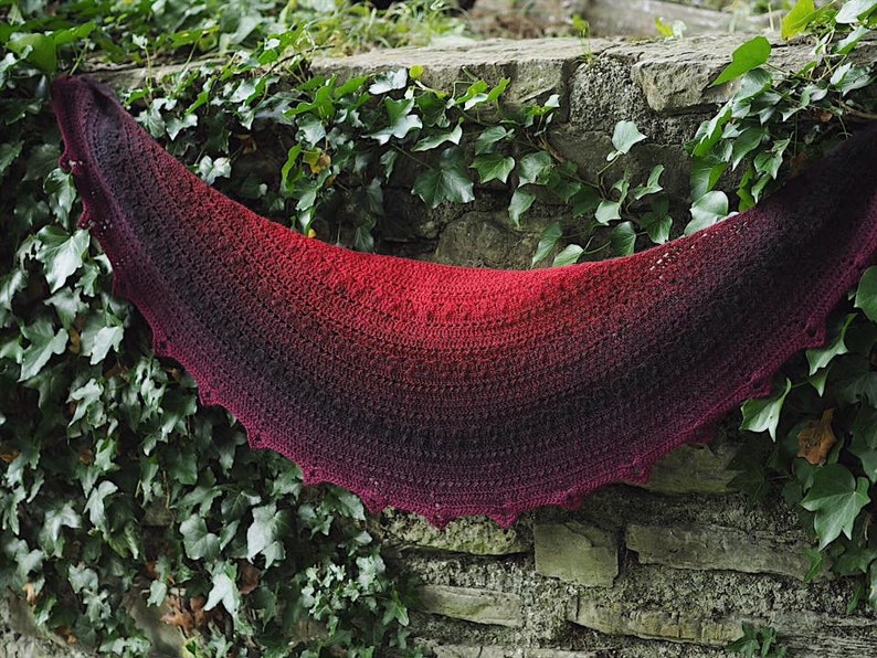 May include: A crocheted shawl with a gradient of red to black. The shawl is draped over a stone wall covered in ivy.