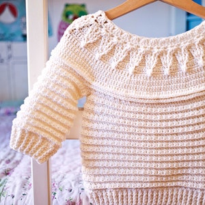 May include: Cream-colored, handmade crochet sweater with long sleeves, hanging on a wooden hanger. The sweater features a textured stitch pattern with a decorative yoke design. The garment is displayed against a soft-focus background, suggesting a cozy, handcrafted aesthetic.