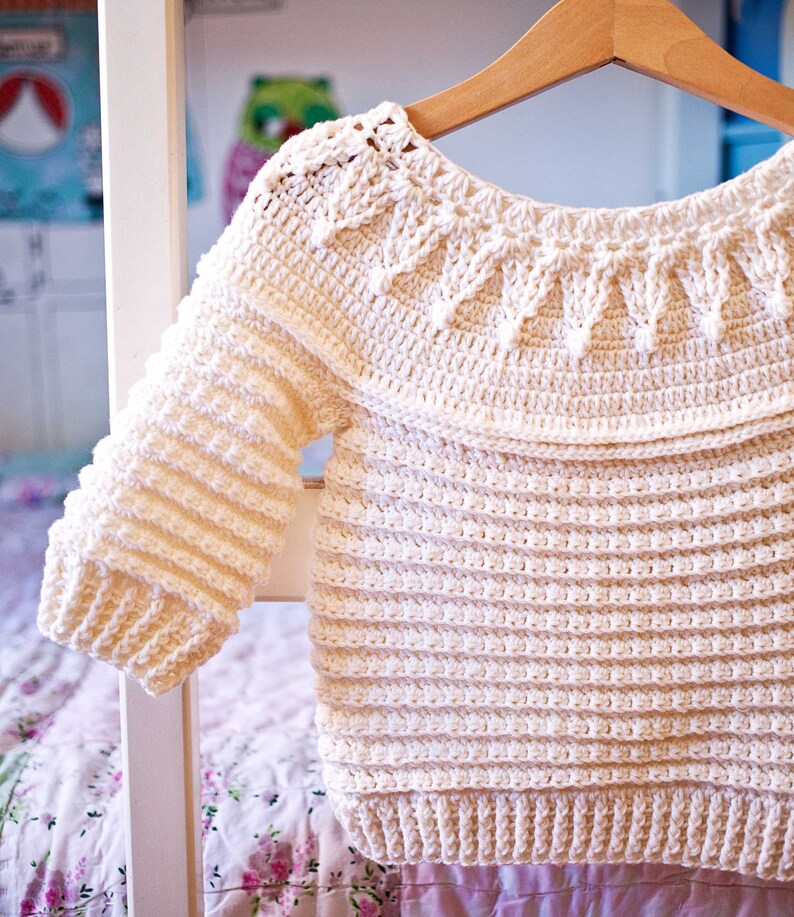 May include: Cream-colored, handmade crochet sweater with long sleeves, hanging on a wooden hanger. The sweater features a textured stitch pattern with a decorative yoke design. The garment is displayed against a soft-focus background, suggesting a cozy, handcrafted aesthetic.