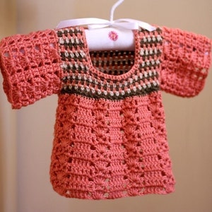 Puede incluir: Un top de bebé tejido a crochet con mangas cortas de color rosa coral. El top tiene un ribete marrón, blanco y beige alrededor del escote.