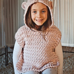 K&ouml;nnte beinhalten: Ein junges M&auml;dchen tr&auml;gt eine rosa gestrickte Kapuzenweste mit B&auml;renohren. Die Weste hat ein strukturiertes Stichmuster und einen gekr&auml;uselten Saum. Das M&auml;dchen tr&auml;gt graue Leggings mit schwarzen Punkten.