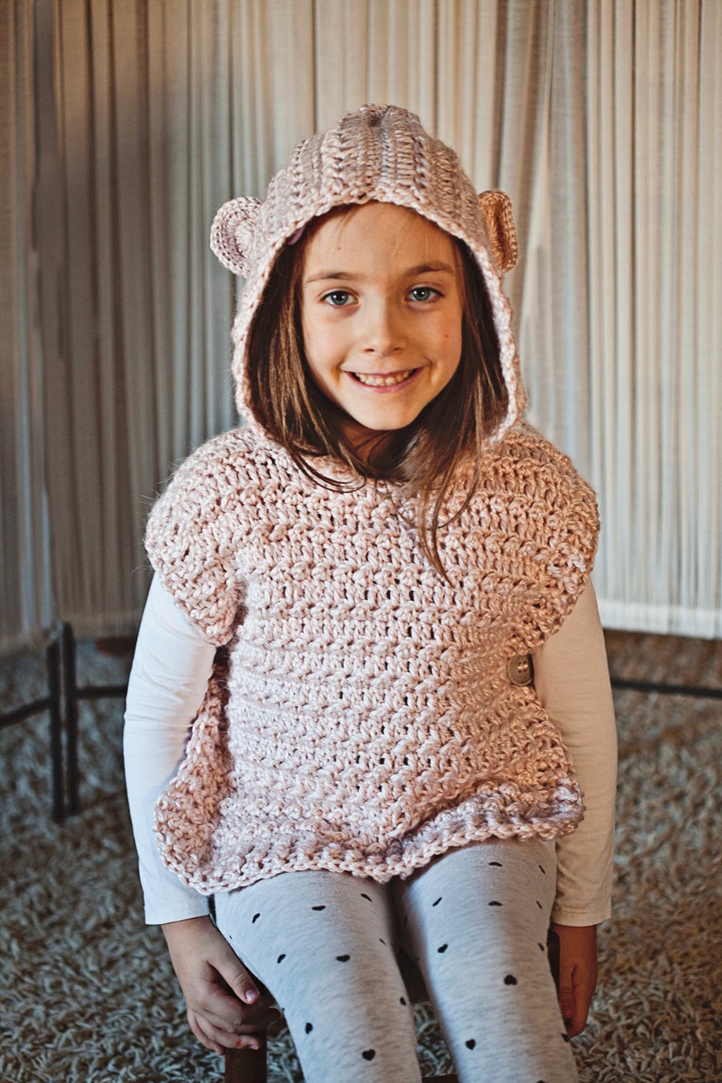 K&ouml;nnte beinhalten: Ein junges M&auml;dchen tr&auml;gt eine rosa gestrickte Kapuzenweste mit B&auml;renohren. Die Weste hat ein strukturiertes Stichmuster und einen gekr&auml;uselten Saum. Das M&auml;dchen tr&auml;gt graue Leggings mit schwarzen Punkten.
