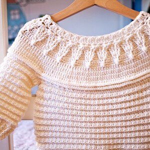 May include: Cream-colored, handmade crochet baby sweater with long sleeves. The sweater features a decorative collar and a textured stitch pattern. It is hanging on a wooden hanger, showcasing the intricate details of the craft.
