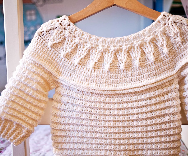 May include: Cream-colored, handmade crochet baby sweater with long sleeves. The sweater features a decorative collar and a textured stitch pattern. It is hanging on a wooden hanger, showcasing the intricate details of the craft.