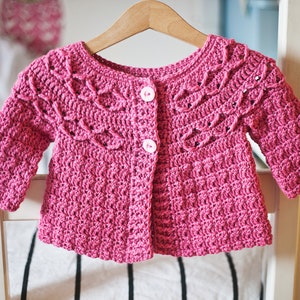 May include: A pink crocheted cardigan with a button closure and a lacy design around the neckline and sleeves.
