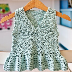 May include: A light green crocheted sleeveless top with a ruffled hem. The top is hanging on a wooden hanger.