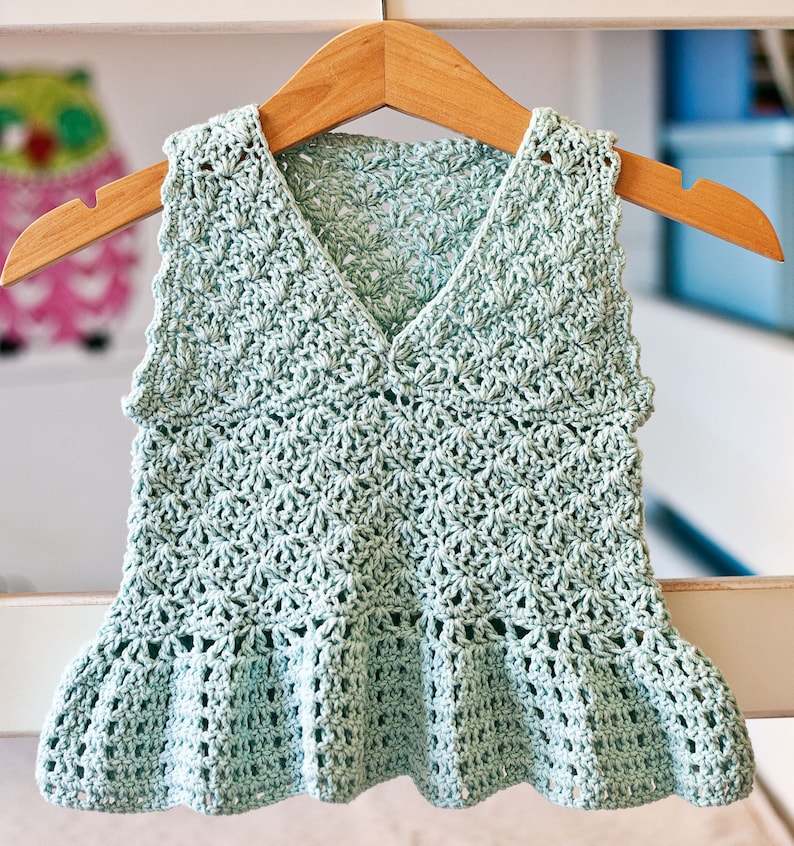 May include: A light green crocheted sleeveless top with a ruffled hem. The top is hanging on a wooden hanger.