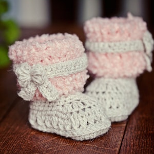 May include: A pair of crocheted baby booties with a pink and gray color scheme. The booties have a fluffy texture and a bow on the front.