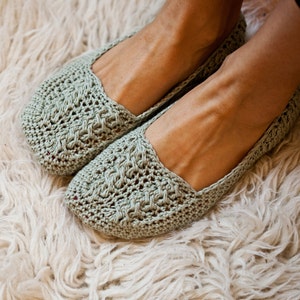 Puede incluir: Un par de zapatillas de crochet verde con un delicado patrón de calado. Las zapatillas se llevan sobre una alfombra blanca y esponjosa.