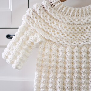 K&ouml;nnte beinhalten: Ein wei&szlig;er H&auml;kelpullover mit einem breiten, gekr&auml;uselten Kragen und einem strukturierten Stichmuster.